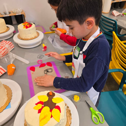 A boy presses out fondant feathers to add to the yellow and red feathers on his fondant turkey atop a white cake.