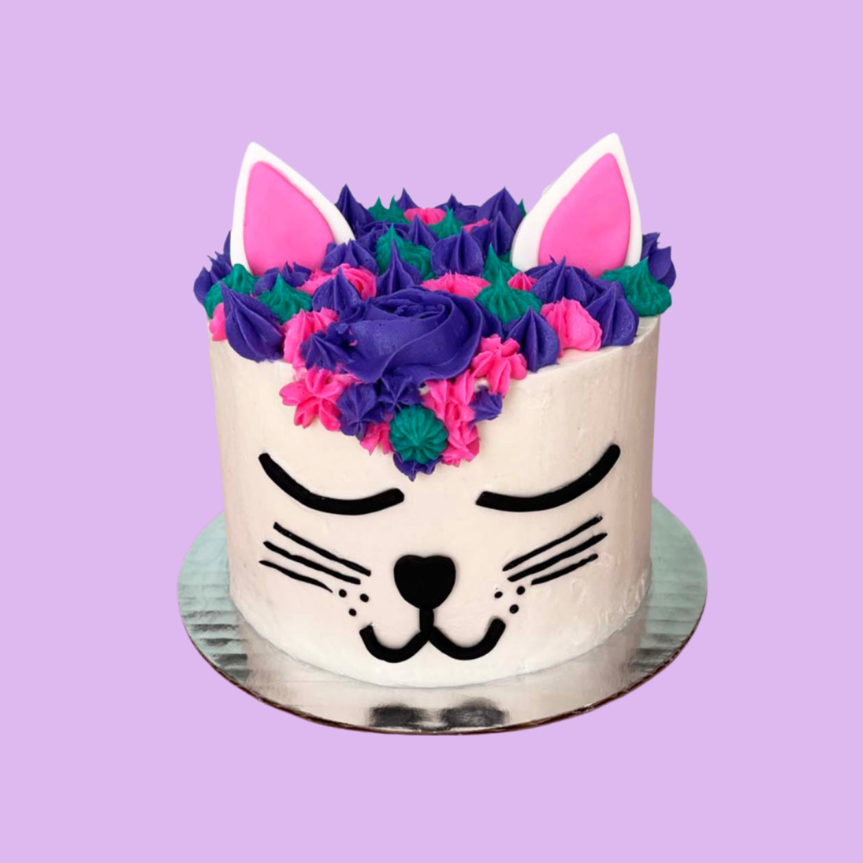 A cat-themed white cake made to look like a cat face with black facial details, purple, teal, and pink frosting hair, and pink and white ears; a DIY cake kit available to decorate during open studio time at Cake Hoopla in Tigard, Oregon, in the Portland area.