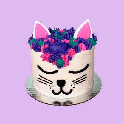 A cat-themed white cake made to look like a cat face with black facial details, purple, teal, and pink frosting hair, and pink and white ears; a DIY cake kit available to decorate during open studio time at Cake Hoopla in Tigard, Oregon, in the Portland area.