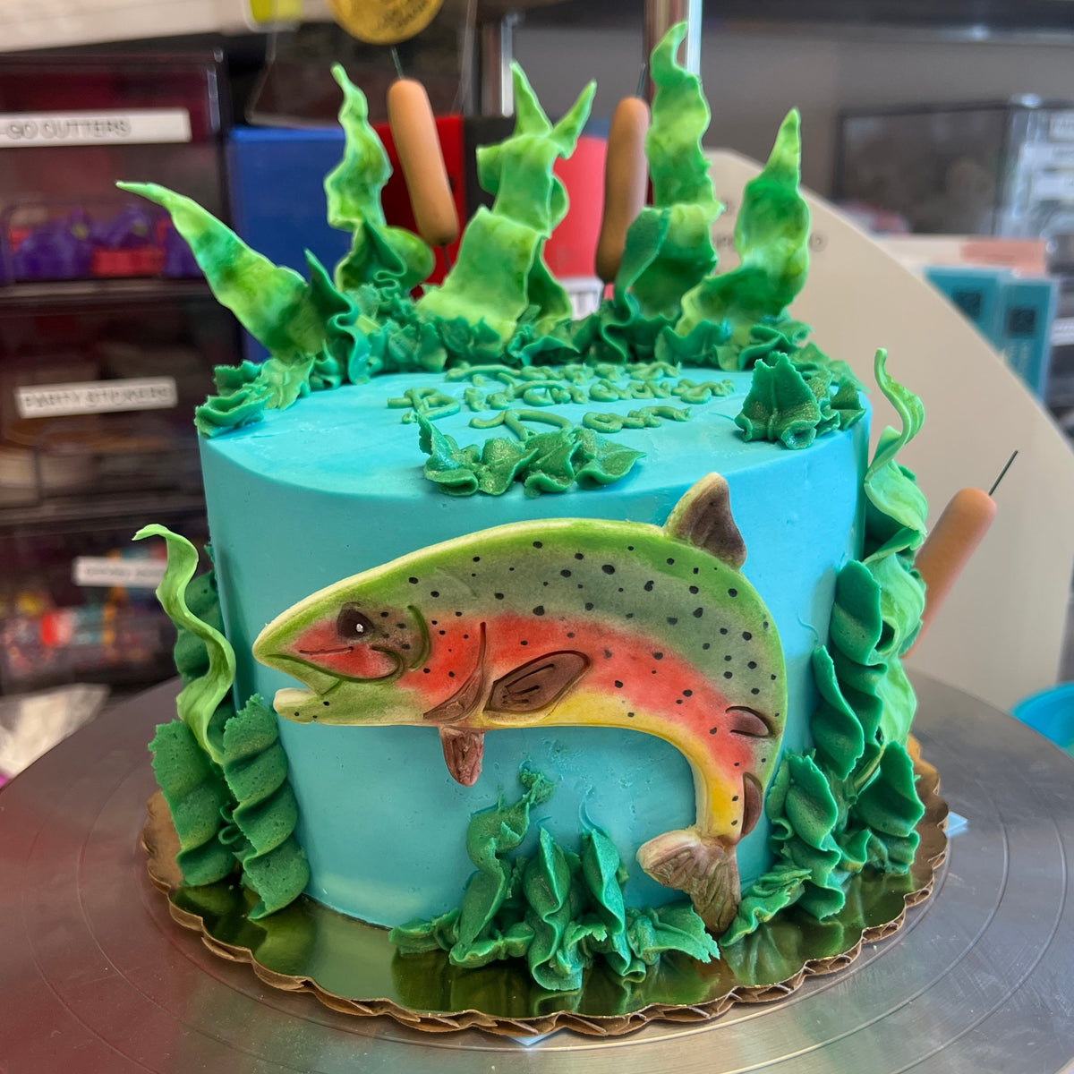 A custom fishing-themed cake decorated with a realistic fondant trout, green iced waterweed, and a bright turquoise blue background, displayed on a silver cake stand.