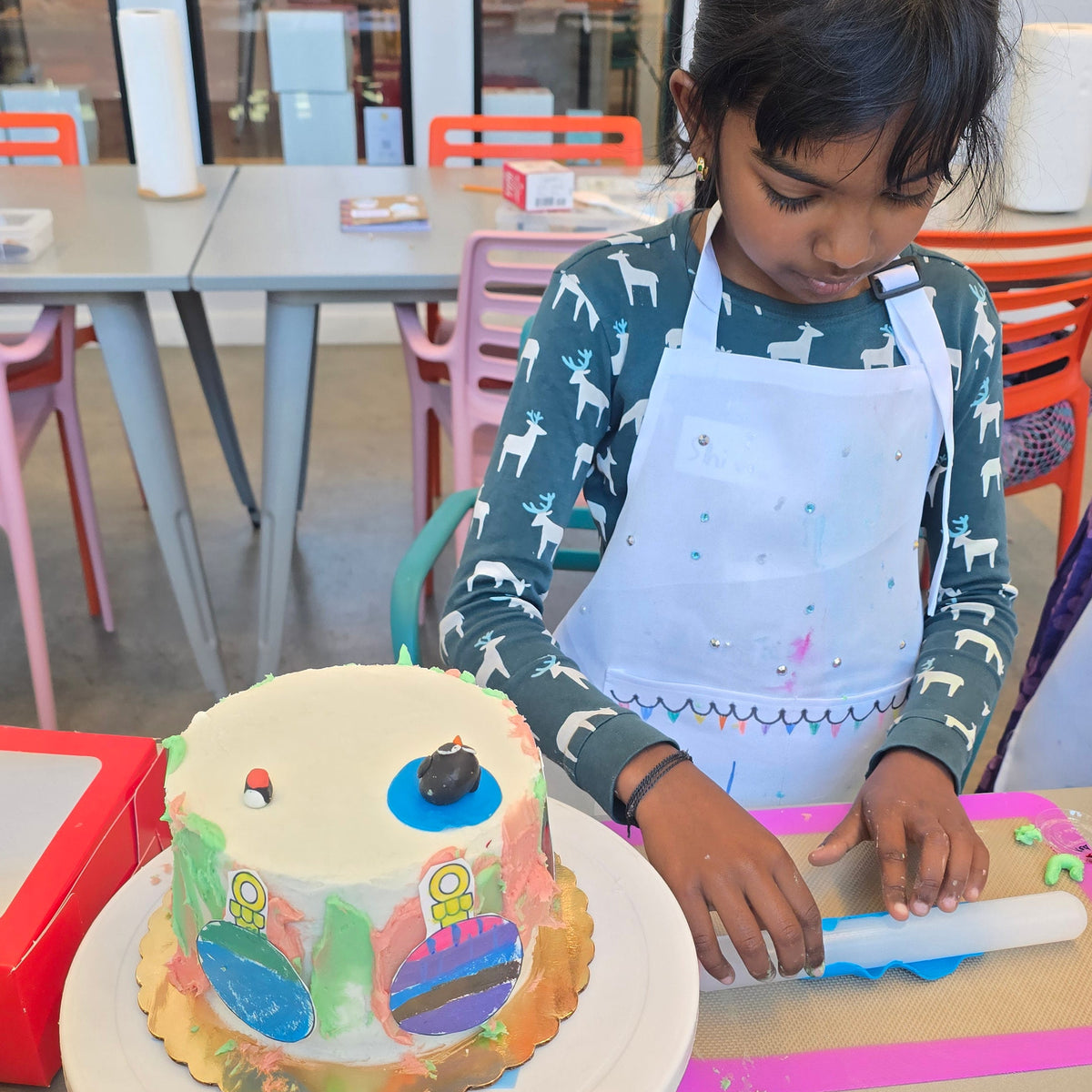 A girl rolls out blue fondant with a rolling pin with her cake just to the side, adorned with painted images of ornaments and a fondant penguin.