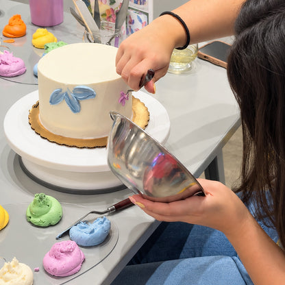 A person's hand holds a bowl and the other a palette knife as they spread on buttercream frosting to form flowers around a whtie cake.