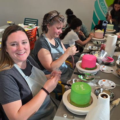 Two women working on their unique cakes with piping bags and colorful frosting at Cake Hoopla's cake-decorating class in Tigard near Portland.