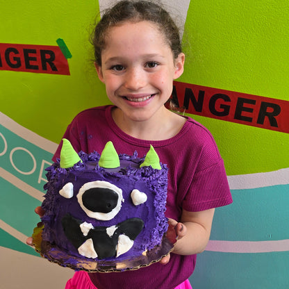 A cake camper shows off her scary and cute 3-horned purple monster with white and black fondant details and green fondant horns.