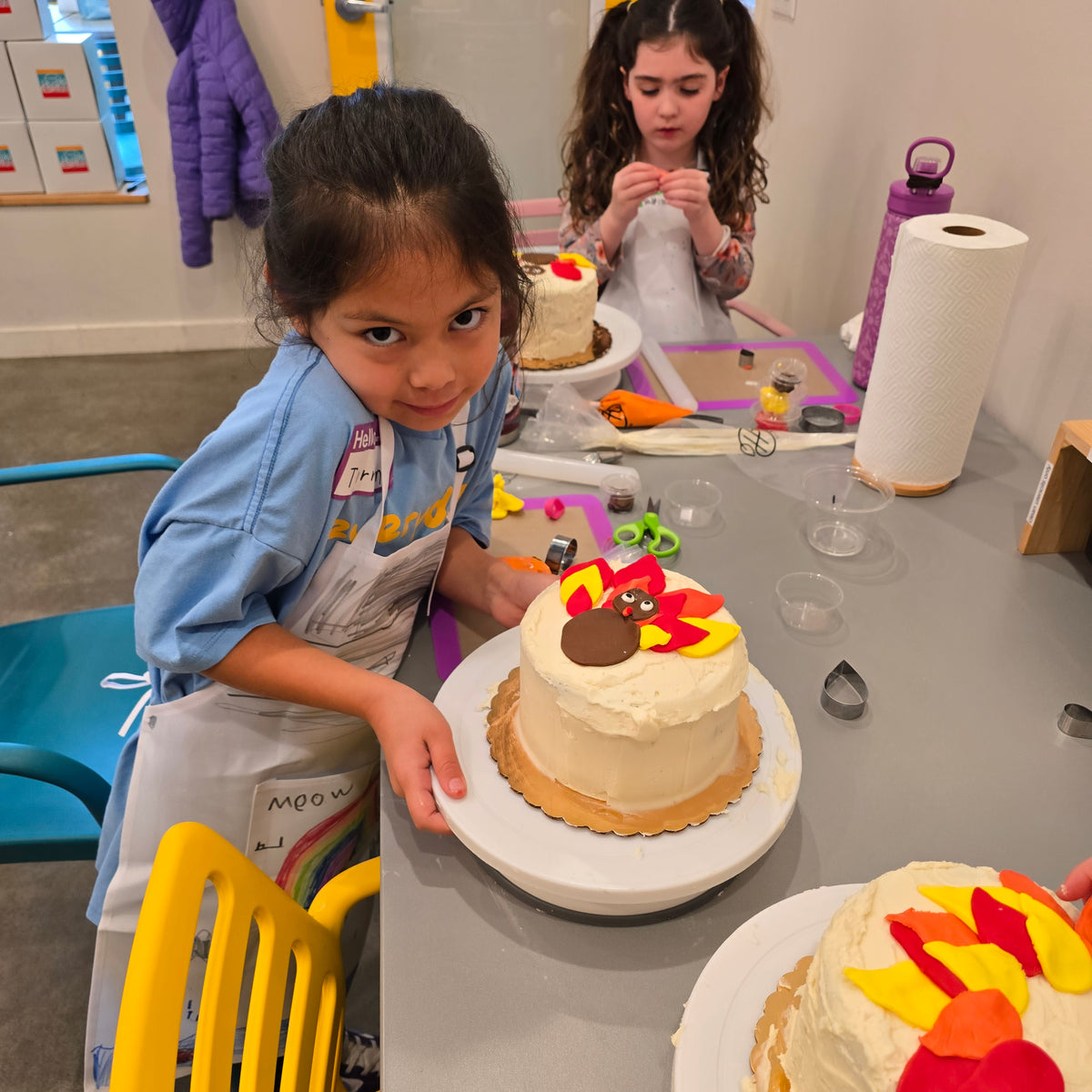 A girl smiles at the camera as she tilts her cake turntable to show off her turkey cake with a brown body and yellow, red, and orange feathers.