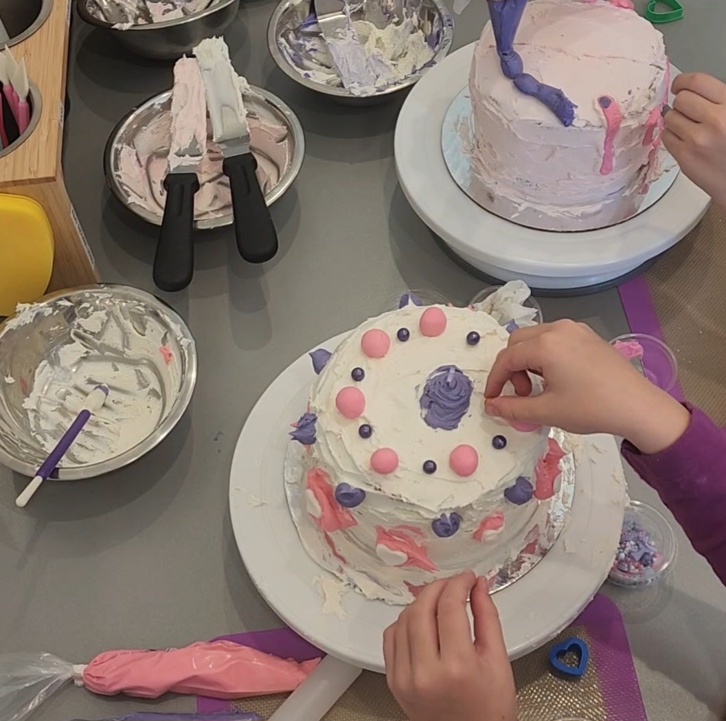 After learning to frost the cakes at the Cake Hoopla DIY cake/cupcake-decorating studio, two kids work on decorating their unique cakes, adding white hearts, purple sprinkles, and pink frosting.