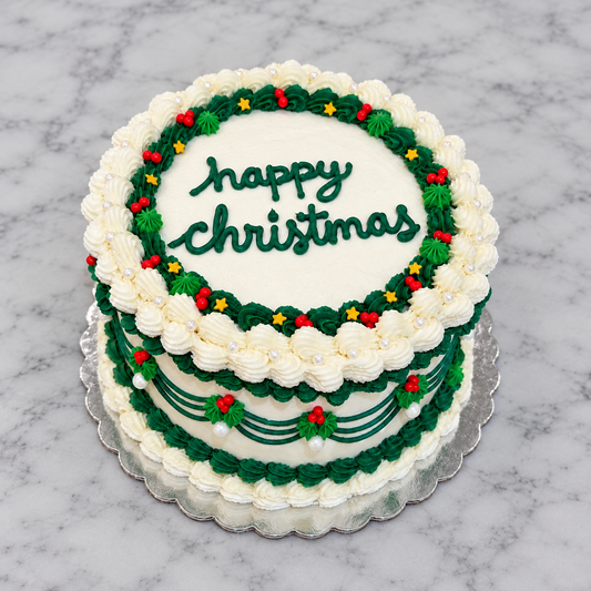 White cake with white and light and dark green frosting borders, red and gold star sprinkles, and "happy christmas" written in dark green frosting on top of the cake.