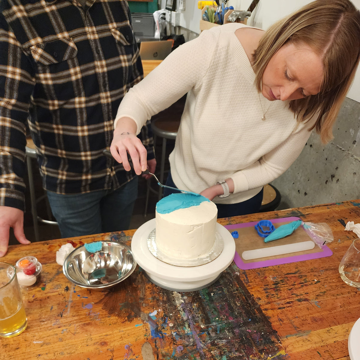 A couple works on frosting a cake together with blue frosting over a white cake.