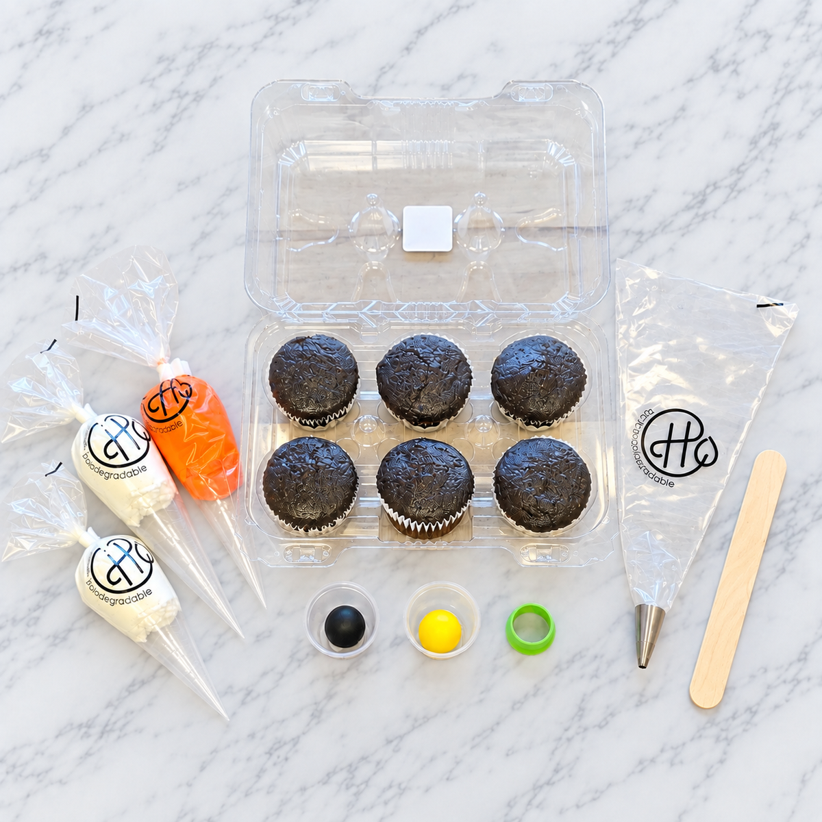 Six chocolate cupcakes surrounded by 2 bags of white frosting, 1 bag of orange frosting, yellow and black fondant, and tools for decorating.