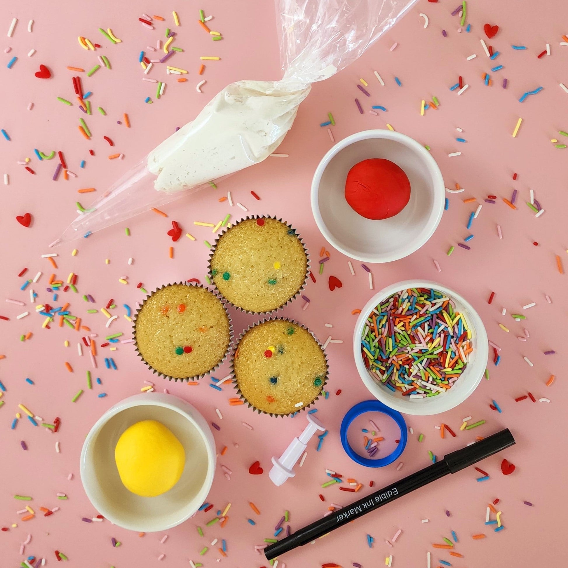 components of Happy Hearts Emoji DIY cupcake-decorating kit, including a piping bag with white frosting, a small heart cutter, a black marker that contains edible ink, a circle fondant cutter, balls of white and red fondant, and rainbow sprinkles. Available during open studio time and to-go from Cake Hoopla in Tigard, Oregon near Portland.