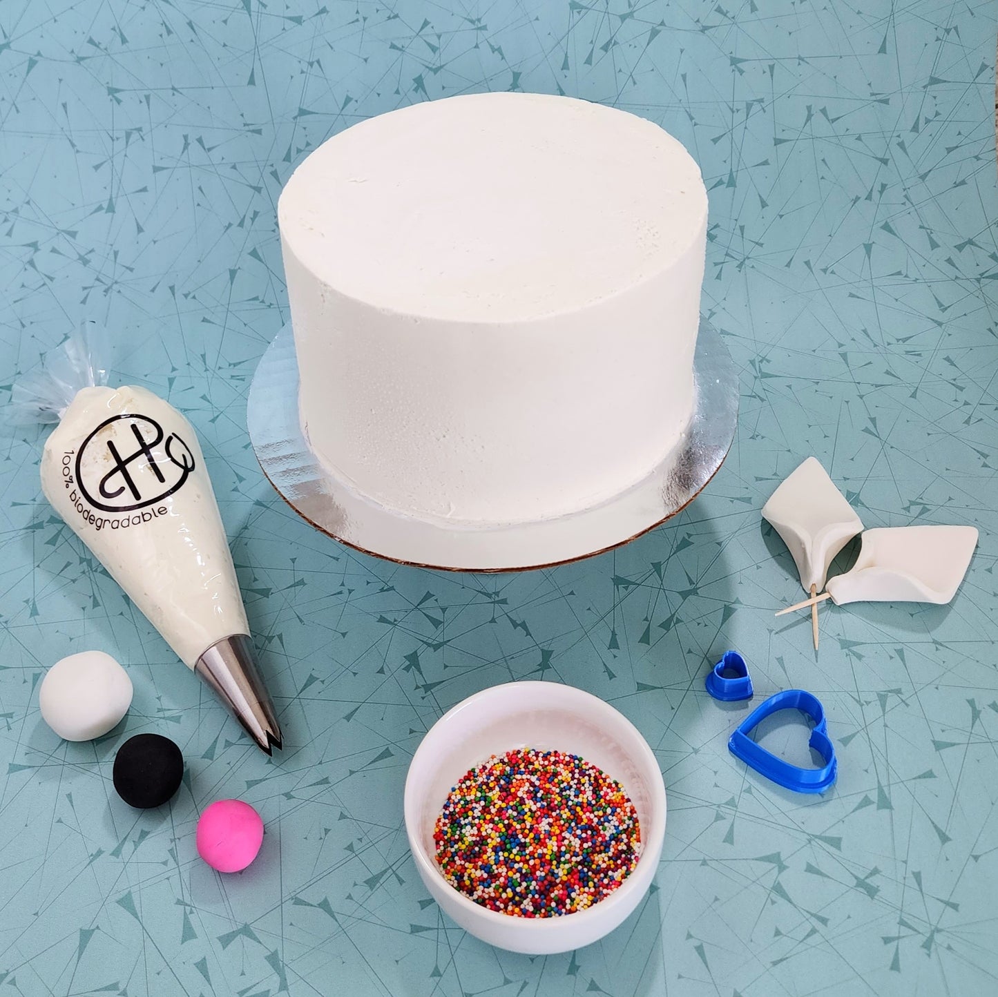 The components of "Shades of Rainbow" DIY cake-decorating kit, including white frosting, white, black, and pink fondant, heart fondant cutters, rainbow sprinkles, and premade white fondant ears.