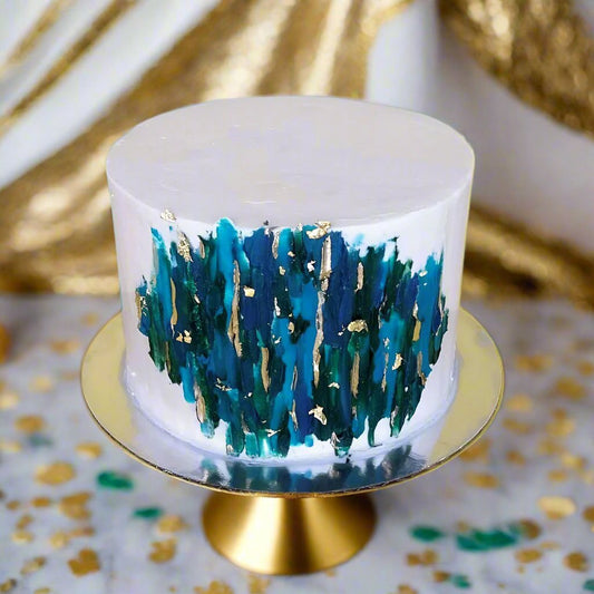 White cake painted with dark green, blue, and teal frosting, gold leaf, and gold edible paint during Cake Hoopla's open studio time in the Portland, Oregon area.