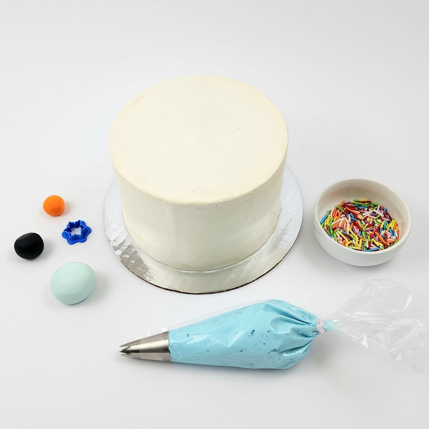 DIY cake decorating kit showing undecorated cake; light blue, black, and orange fondant; blue frosting with piping tip; snowflake cutter; and rainbow sprinkles.