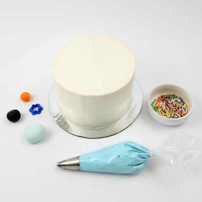 DIY cake decorating kit showing undecorated cake; light blue, black, and orange fondant; blue frosting with piping tip; snowflake cutter; and rainbow sprinkles.