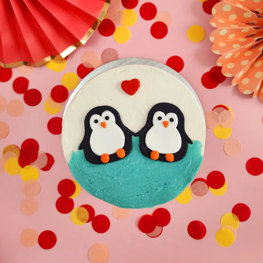 Two fondant penguins jumping into blue frosting with a red heart floating above. Finished I Flippin' Love You DIY cake decorating kit.