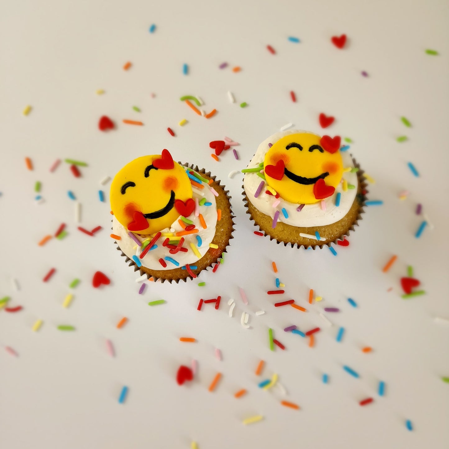 Emoji cupcakes frosted white with rainbow sprinkles and yellow smiling emoji faces with pink cheeks and three floating hearts; pre-decorated cupcakes available for pick up at Cake Hoopla in Tigard, Oregon, in the Portland area.