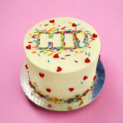 A Just Hi! DIY cake with the word "Hi!" written out of rainbow sprinkles and fondant red hearts dotting the sides, available as a DIY cake-decorating kit during open studio time or to-go or as a pre-decorated cake at Cake Hoopla in Tigard, Oregon near Portland.