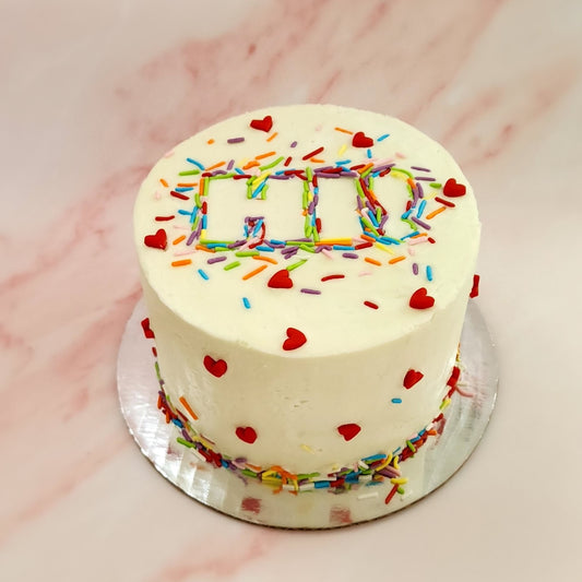 A decorated DIY Just Hi cake. White frosted cake decorated with rainbow sprinkles forming the word "Hi" in the center and cute red fondant hearts dotted around the front and sides of the cake.