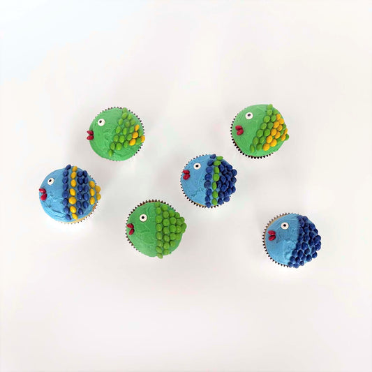 Several finished DIY Keep It Reel cupcakes look like fish. They are frosted blue and green and have scales made out of blue, green, and yellow chocolate candies. Each fish has a fondant googly eye and red lips made from chocolate candies. A unique activity for an under-the-sea or ocean-themed party or event.