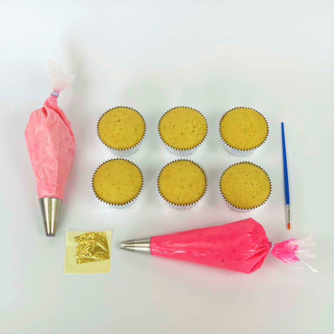 The components of the DIY La Fleur cupcake kit, including gold leaf, a paintbrush, six of the 12 cupcakes that come with the kit, and light pink and dark pink frosting in piping bags, each with a different silver piping tip.