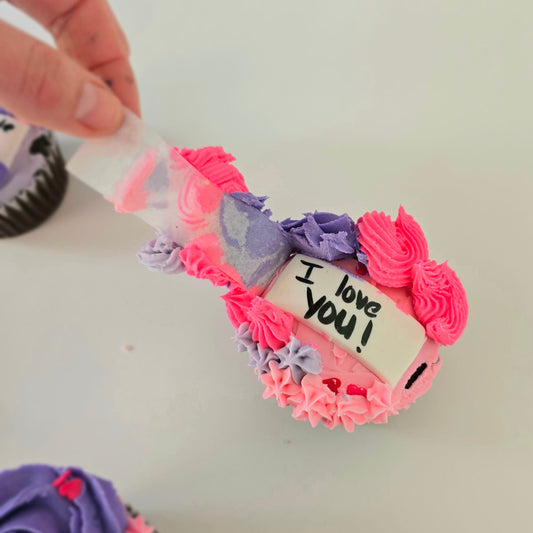 Hidden message cupcakes decorated with purple and pink frosting with stars and rosettes as a hand peels back parchment to reveal the hidden message, "I love you!" during Cake Hoopla's Hidden Message Valentine's cupcake-decorating class in Tigard, Oregon near Portland.