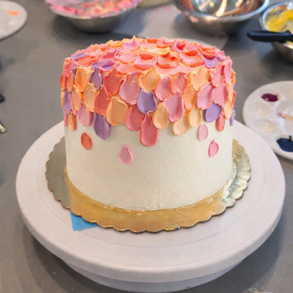 Colorful cake with pink, orange, and purple spatula decorations on a white stand amongst cake-decorating tools.