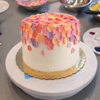 Colorful cake with pink, orange, and purple spatula decorations on a white stand amongst cake-decorating tools.
