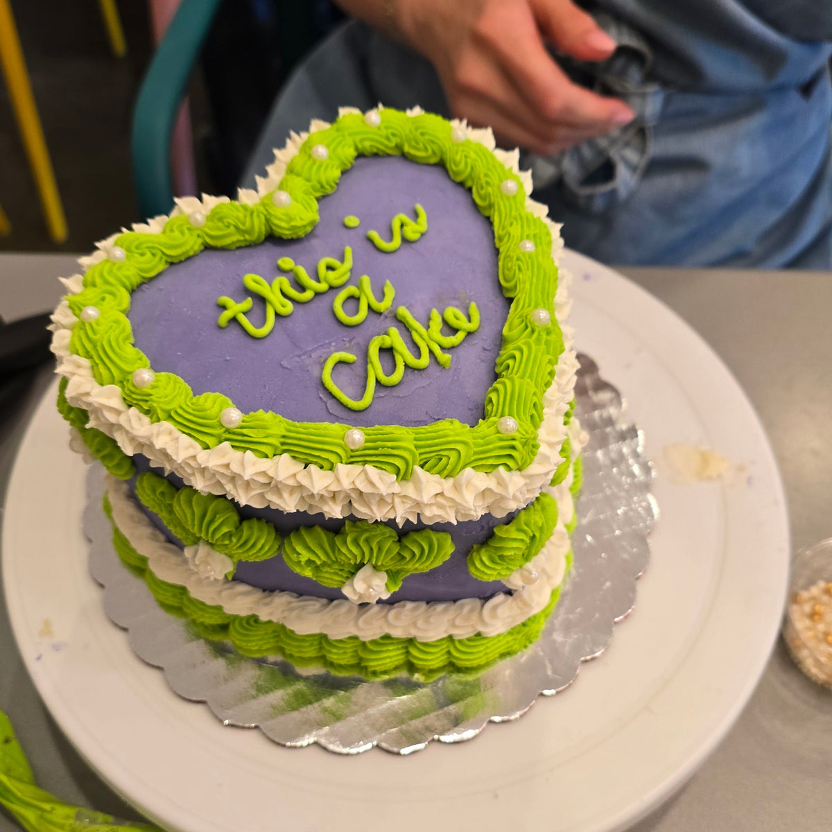 Vintage heart-shaped cake with green and purple icing on a white turn table created by a student at Cake Hoopla's cake-decorating class.