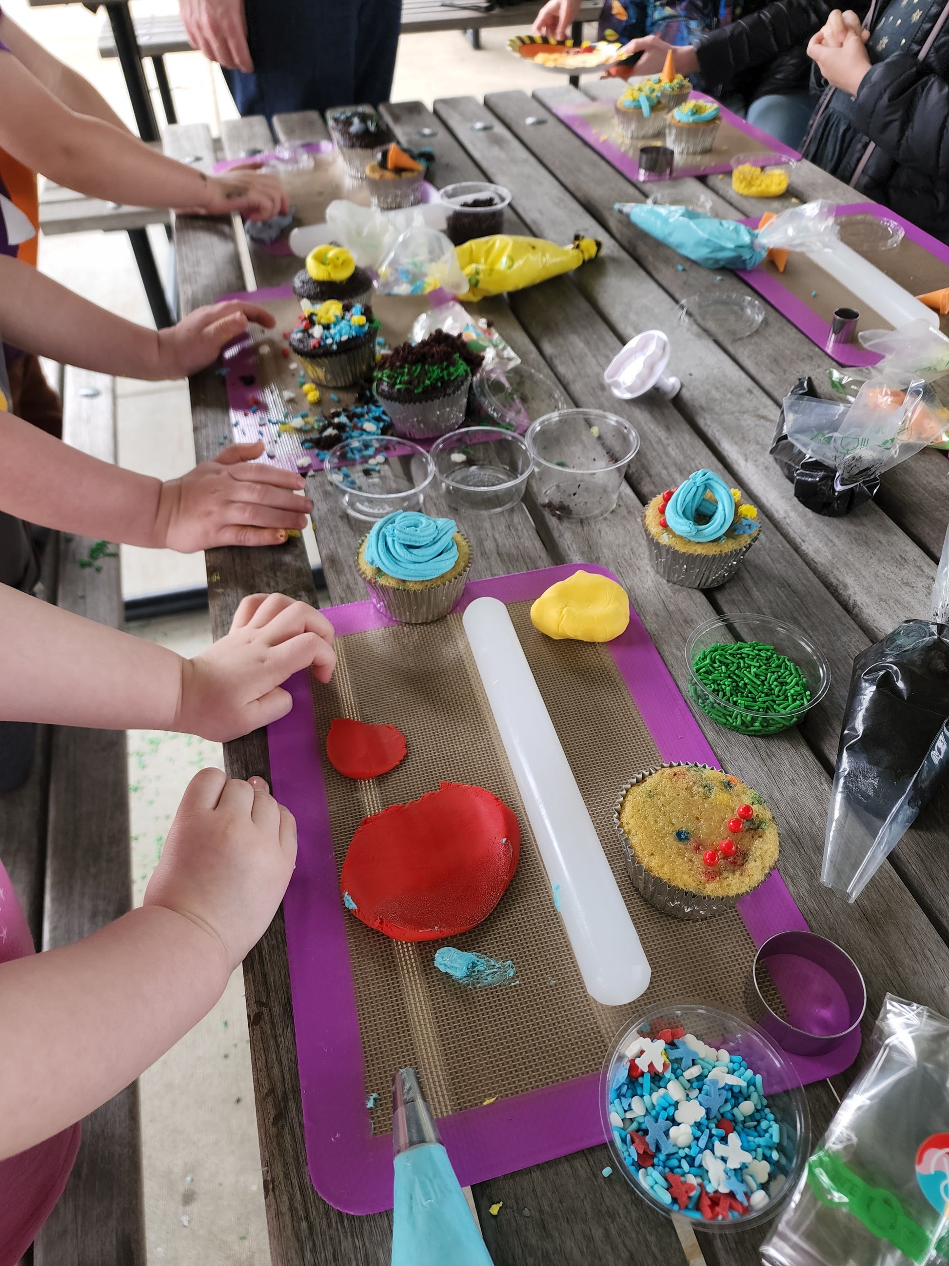 The scene of active little hands decorating cupcakes at a custom cake party in the Portland area. A picnic table is topped with a smattering of cupcake-decorating ingredients and tools, like fondant, rollers, cutters, frosting, sprinkles, and edible dirt.