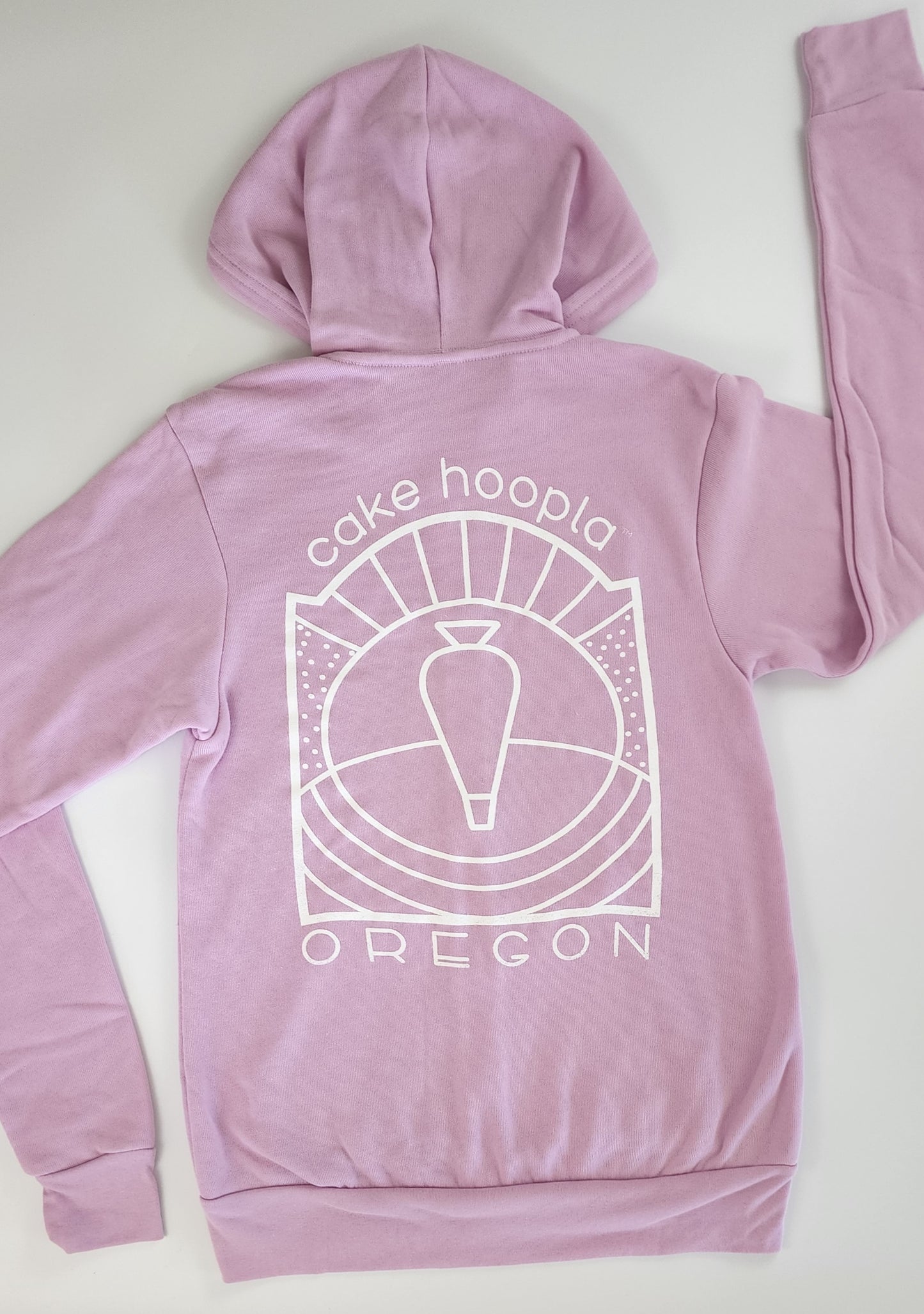 The backside of a unisex light-purple zip-up hoodie. The white screen printing in the center of the hoodie forms an abstract outlined image of a piping bag in front of a hill with rays of sunshine bursting from the center. Above the piping bag artwork is "cake hoopla" written in simple white lettering. Under the artwork is "OREGON" in caps with a slightly retro feel. The artwork combined with the nod to Oregon depicts Oregon's wine country.