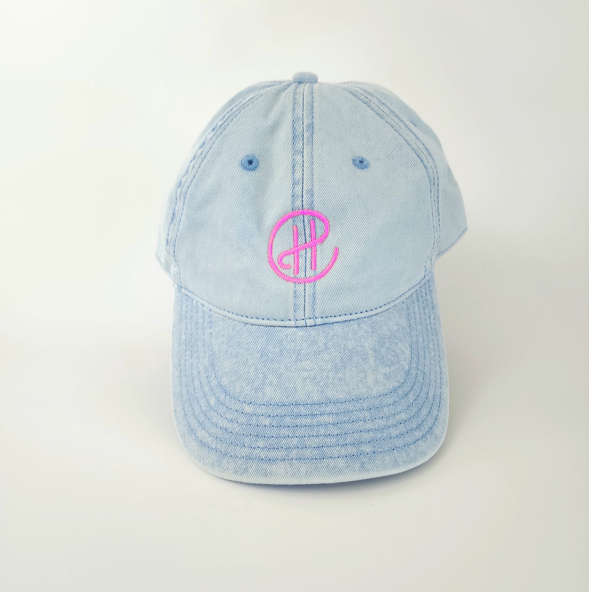 The front of a light-blue denim hat with a neon pink "CH" embroidered in the center. In the CH logo, the center bar of the H connects to the C, which encircles the H. The logo stands out bright against the blue denim. This hat has a ‘90s retro feel contrasted with modern-looking embroidery.