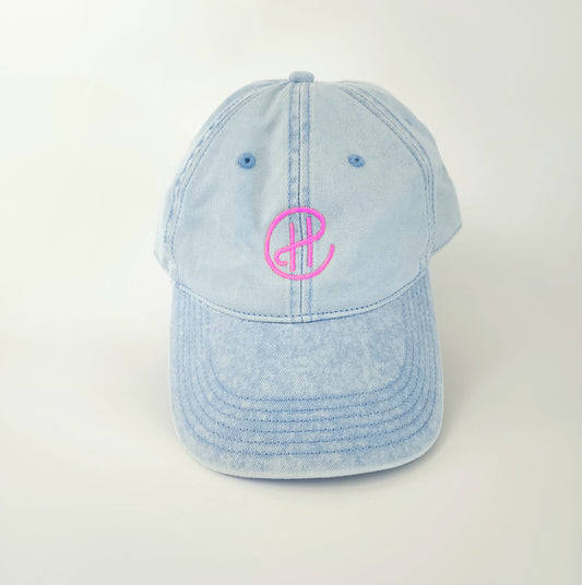 The front of a light-blue denim hat with a neon pink "CH" embroidered in the center. In the CH logo, the center bar of the H connects to the C, which encircles the H. The logo stands out bright against the blue denim. This hat has a ‘90s retro feel contrasted with modern-looking embroidery.