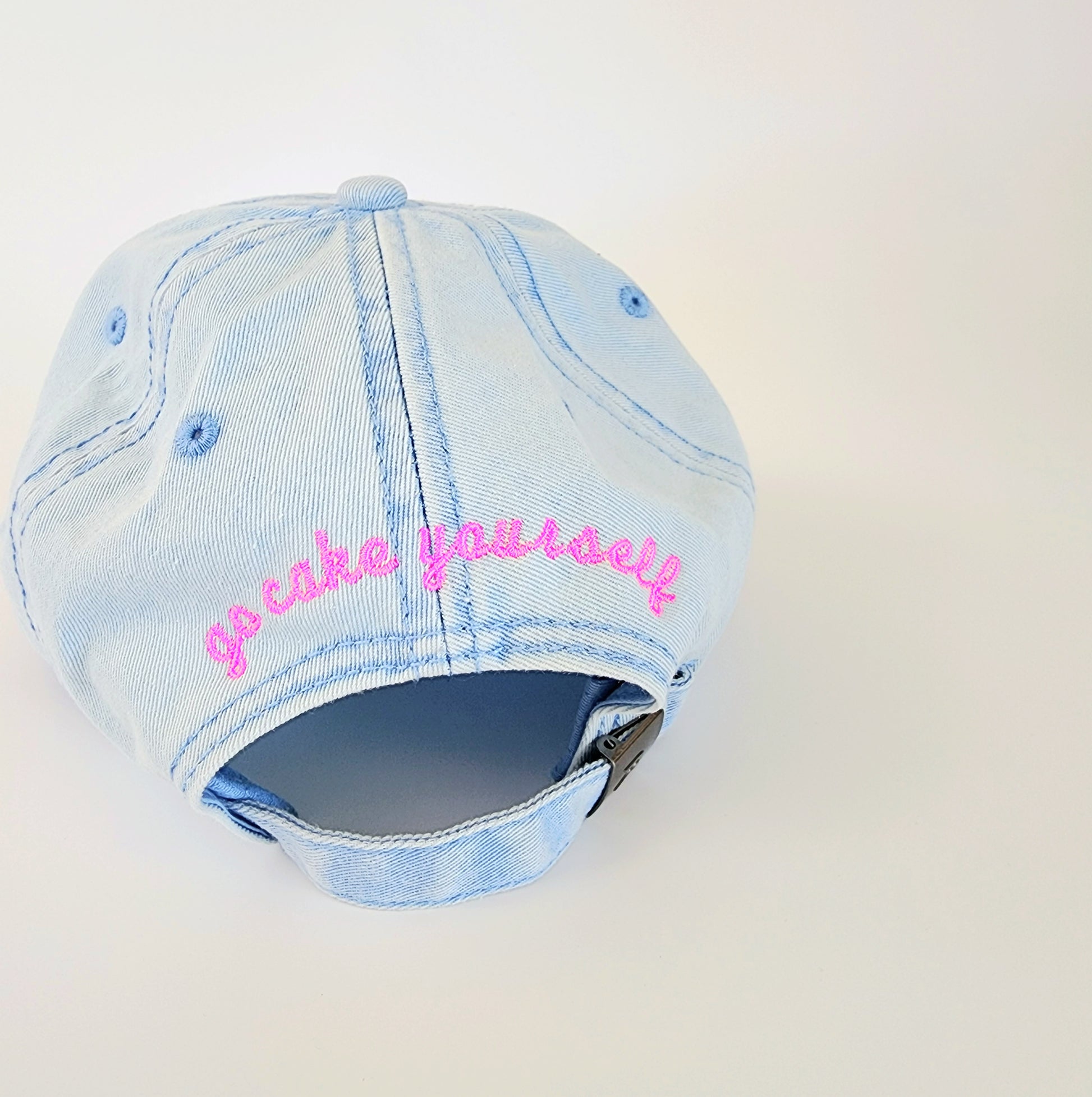 The back of a light-blue denim hat with an adjustable strap to make the hat bigger or smaller. In an arch above the adjustable strap are the words "go cake yourself" in bright pink lowercase cursive. The adjustable strap is fastened by a brass-colored lift-type buckle. This hat has a ‘90s retro feel contrasted with modern-looking font.