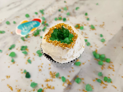 A closeup of one of the finished geode cupcakes from the You’re a Gem cupcake kit. White vanilla buttercream frosting glistens around gold sparkly sprinkles around green rock candy that forms a geode shape in the middle of the cupcake.