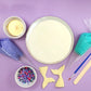 A colorful array of the tools and ingredients you receive with this DIY mermaid cake kit: gold paint, purple and teal frosting in piping bags, a paintbrush, a wood spatula, two white chocolate mermaid tales, sprinkle mix with purple, pink, white, blue, and gray sprinkles, and a cake frosted in white vanilla buttercream. Not shown is the easy-to-follow instruction card you will also receive.