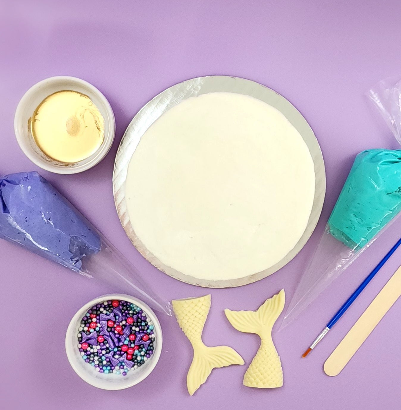 A colorful array of the tools and ingredients you receive with this DIY mermaid cake kit: gold paint, purple and teal frosting in piping bags, a paintbrush, a wood spatula, two white chocolate mermaid tales, sprinkle mix with purple, pink, white, blue, and gray sprinkles, and a cake frosted in white vanilla buttercream. Not shown is the easy-to-follow instruction card you will also receive.