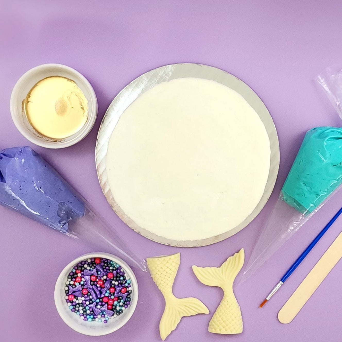 The components of the "Mermaid's Tale" DIY cake-decorating kit: gold paint, purple and teal frosting in piping bags, a paintbrush, a wood spatula, two white chocolate mermaid tales, sprinkle mix with purple, pink, white, blue, and gray sprinkles, and a cake frosted in white vanilla buttercream. Not shown is the easy-to-follow instruction card you will also receive.