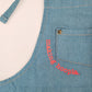 A closeup view of the corner of the breast pocket outlined with neon pink embroidery reading "making hoopla." The font has a typewritten look and contrasts nicely against the blue of the apron. Kids will stay clean and stylish in this apron.