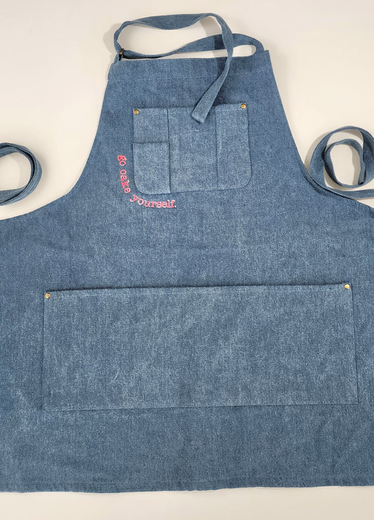 A medium-blue denim apron with a utility pocket across the breast. Tracing the bottom corner of the pocket are the words "go cake yourself" in neon pink embroidery. Across the lower body of the apron is a large open pocket. Small bras rivets stamp the upper corners of each pocket. A denim neck strap is secured with a brass-colored loop buckle. 2 long straps rest on either side of the apron. This apron is modern with denim a slight Americana feel. 