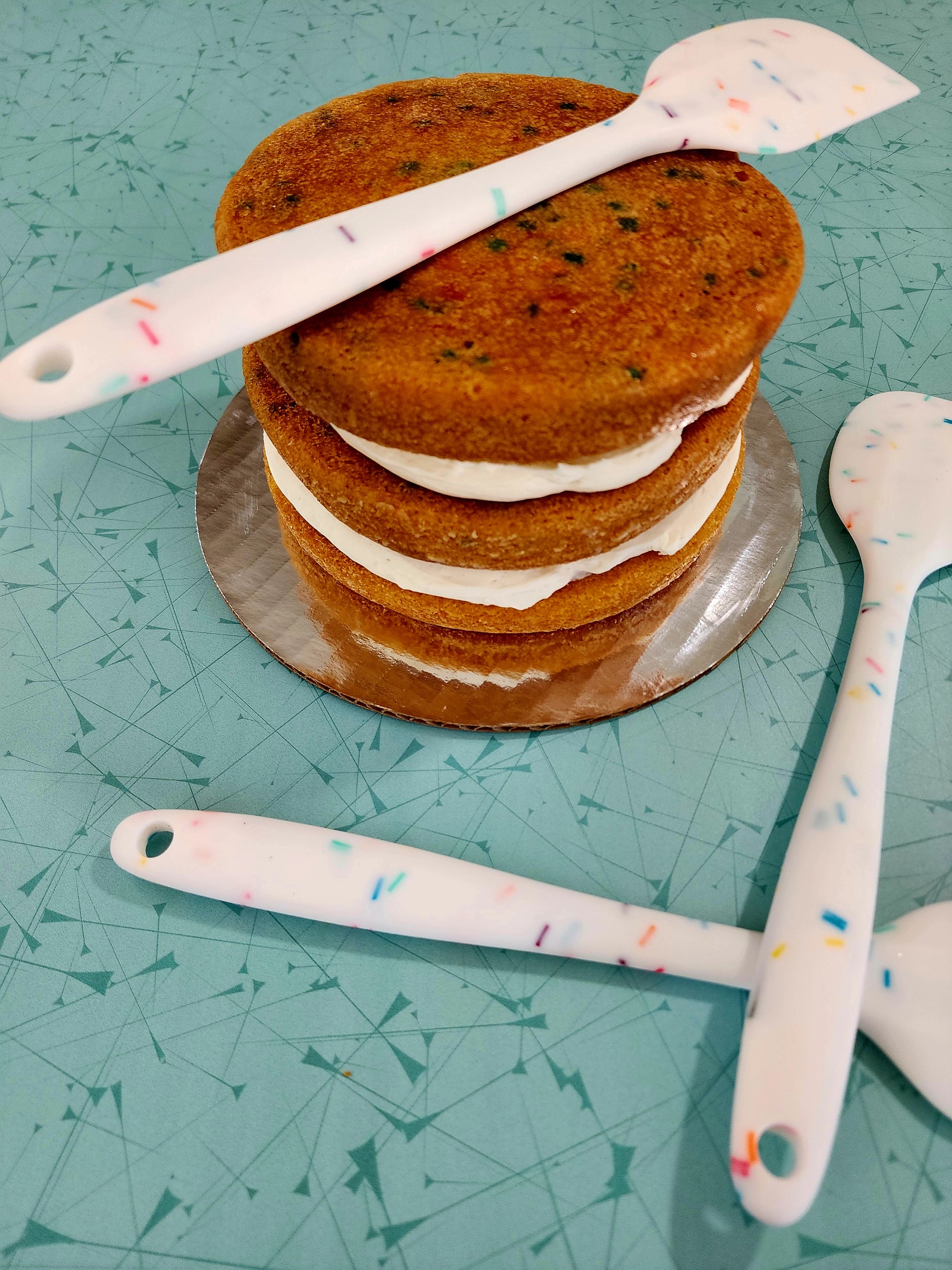 Several silicone spatulas lay on top of and around an unfrosted three-layer Fetti cake. These spatulas would be handy for frosting the cake. 