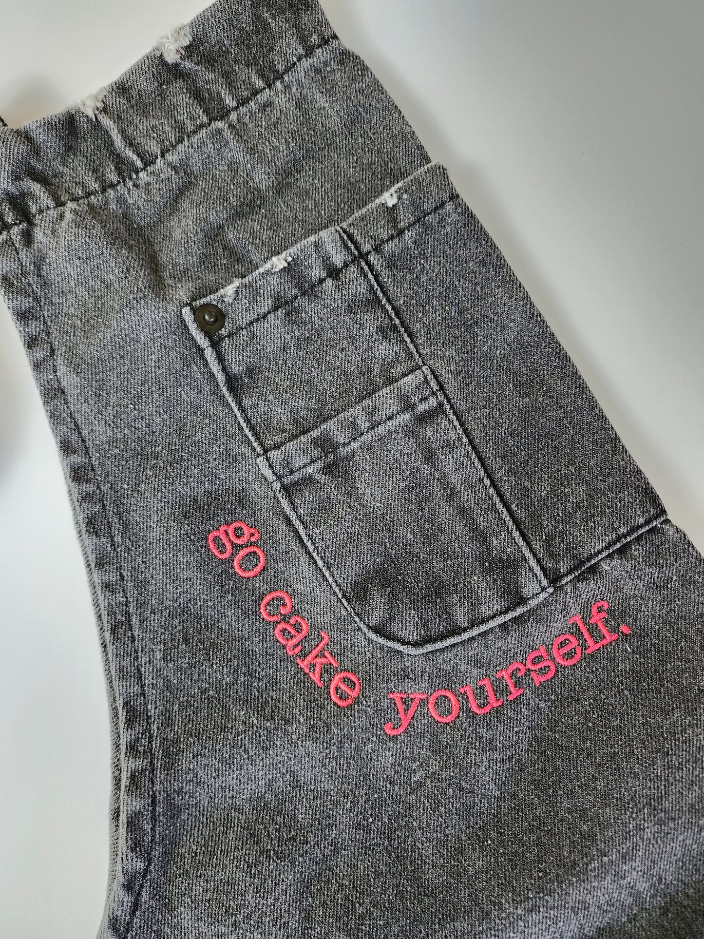 A closeup view of the corner of the breast pocket outlined with neon pink embroidery reading "go cake yourself." The font has a typewritten look and is somewhat discrete for the size of the apron. 