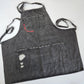 A black distressed denim apron has a utility pocket at the breast and a large open pocket at the bottom. Partially outlining the breast pocket are the words "go cake yourself" in neon pink embroidery. The bottom pocket has extra distressing in one corner for an almost torn look. Long thin straps are attached to either side of the apron, as well as a neck strap at the top secured by a brass-colored loop buckle. This apron has a classic look with punk details.