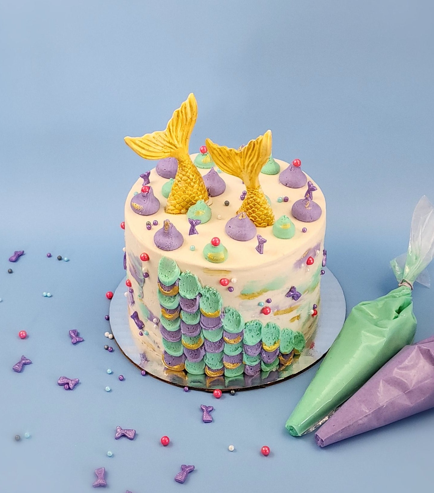 The finished DIY mermaid cake decorated with pink, purple, blue, white, and gray sprinkles, purple mermaid tales, and two large gold mermaid tales sticking out of the top. Two piping bags, one with teal and one with purple frosting, sit next to the cake, surrounded by sprinkles on a blue background.