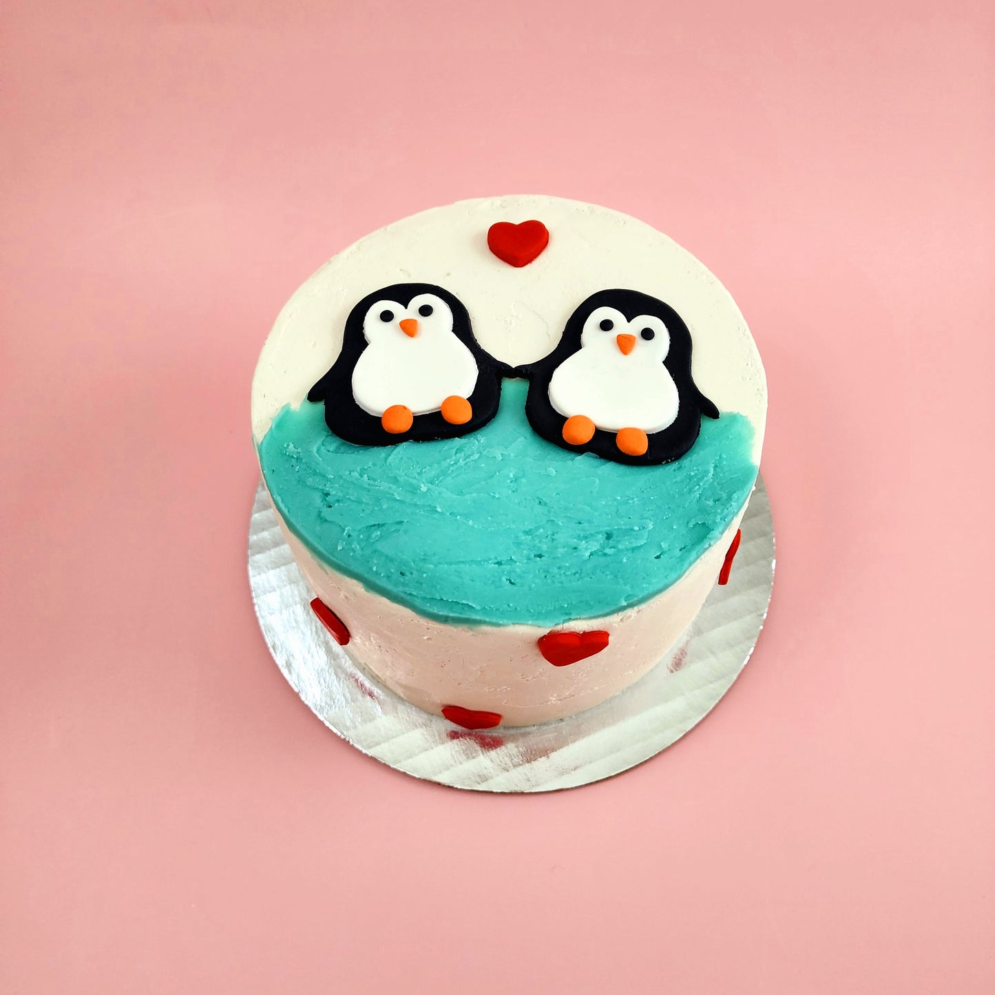 A love-themed white cake with teal blue frosting and two black, white, and orange penguins jumping into the water and red fondant hearts dotting the cake; a pre-decorated cake available for pick up at Cake Hoopla in Tigard, Oregon, in the Portland area.