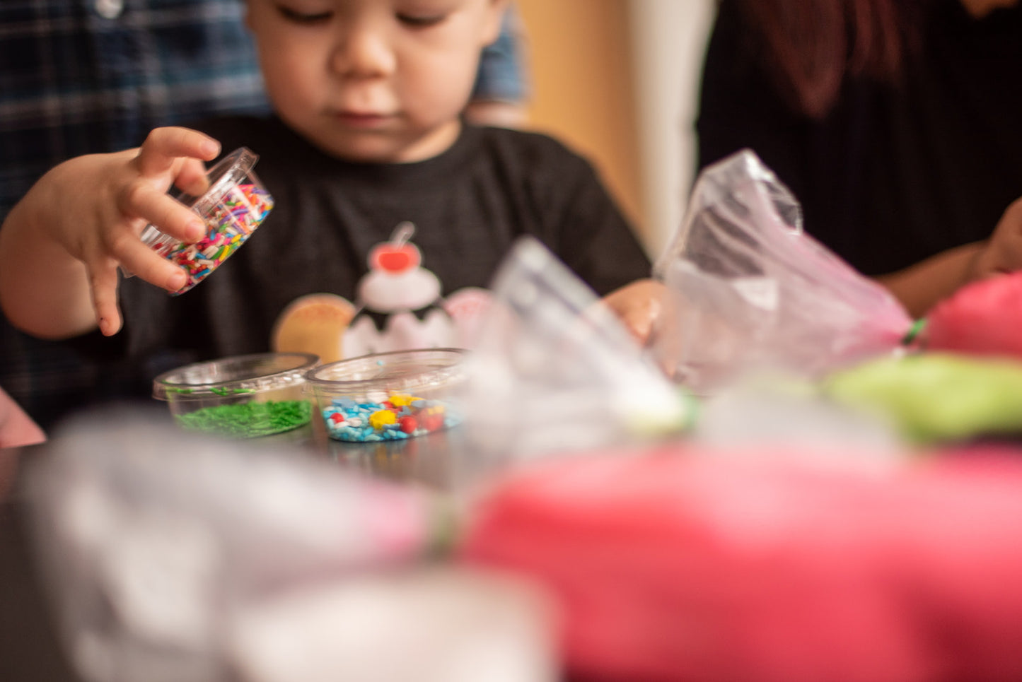 A close-up picture of a little boy at a custom DIY cake party in the Portland area. The boy holds a container of sprinkles. Other cups of sprinkles and piping bags of colorful frosting rest on the table in front of him.