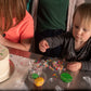 A little girl and a toddler are at a custom cake decorating party in the Portland area. They are decorating a white cake with pink frosting, fondant, and several colors of sprinkles. 