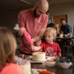 At a custom DIY cake decorating party in the Portland area, a father and son decorate a white cake with pink frosting from a piping bag. They are both concentrating and appear happy. Other groups of kids and parents are also decorating cakes. 