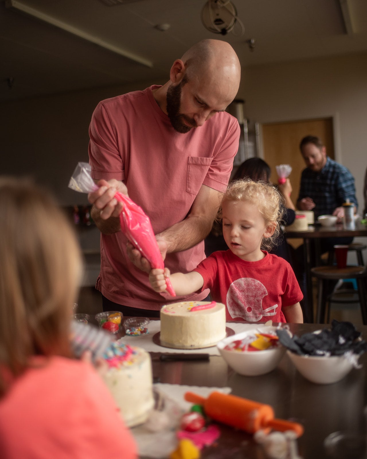 At a custom DIY cake decorating party in the Portland area, a father and son decorate a white cake with pink frosting from a piping bag. They are both concentrating and appear happy. Other groups of kids and parents are also decorating cakes. 