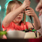 A little girl is at a DIY cake decorating party in the Portland area and is concentrating on piping frosting onto the top of her cake, where a mound of frosting and sprinkles already exists. Another out of focus kid is decorating their cake with a spatula. 