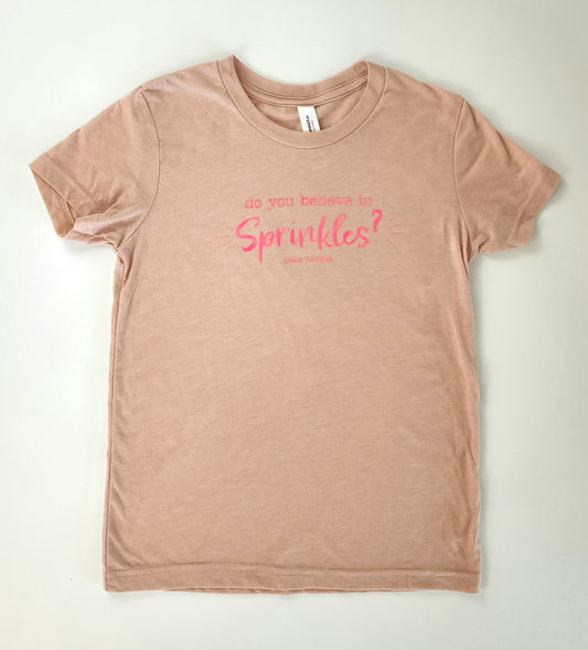 A stylish and kid-friendly peachy-mauve-colored short-sleeved t-shirt. In the center of the t-shirt in neon pink writing is the question "do you believe in Sprinkles?" The font for "sprinkles" is a whimsical cursive-style font, contrasted against the typewriter-style font of the words "do you believe in.” Beneath this phrase are the words "cake hoopla" in small neon-pink print.